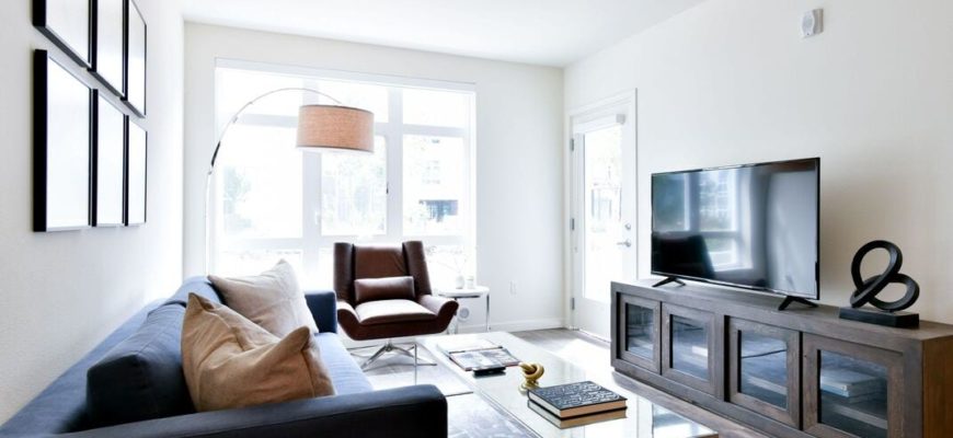 living room with blue couch on the left, tv stand and tv on the left, wall art on the right wall and big open windows in the background.